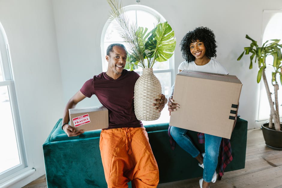 A man and a woman standing inside a doorway, each holding cardboard boxes during a home relocation process. The man on the left has dark skin, short black hair, and is wearing a dark red shirt with orange pants, carrying a large cardboard box. The woman on the right has light skin, long brown hair, and is dressed in a black and white checkered shirt with beige pants, holding a smaller box labeled 'FRAGILE' with red tape. They are positioned in a narrow corridor with plain white walls and a dark door frame behind them. The scene suggests a packing and moving activity, with the individuals preparing to transport household items, illustrating the loading process typical of professional removals services like those offered by Removals Kingston upon Thames. The environment is well-lit, emphasizing the details of the packaging materials and the cautious handling of the boxes.