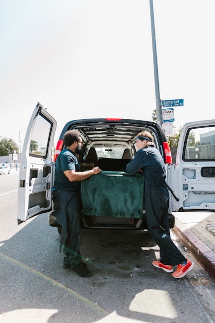 Two movers from Removals Kingston upon Thames are loading a large, dark green upholstered piece of furniture into the open back of a white commercial van, which is parked on a paved street. The furniture is partially wrapped in protective fabric, indicating careful handling during the packing and moving process. One mover is wearing a dark uniform with safety glasses, while the other is dressed similarly, both working together to lift and position the furniture inside the vehicle. The van's interior reveals minimal shelving or objects, focusing on the furniture being prepared for home relocation. The surrounding environment shows a clear, sunny day with a street sign visible that reads 'Somerset.' The scene highlights the practical steps involved in furniture transport, typical of professional relocation services like those offered by Removals Kingston upon Thames, and demonstrates the loading process during a house move or collection from a property located near Canbury Gardens.