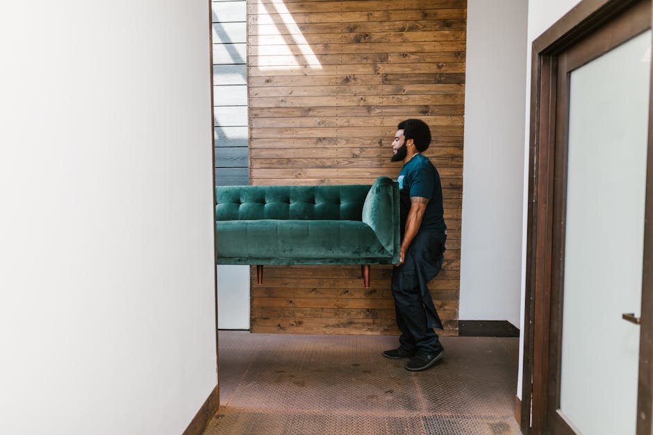 A man from Removals Kingston upon Thames is engaged in a furniture transport process inside a building, lifting a green velvet sofa with wooden legs. The sofa, partially wrapped in clear plastic, is being moved through a doorway into or out of a room with a wooden panel wall in the background. The interior features neutral decorated walls with a brown carpeted floor, and the man is wearing a dark blue uniform with black shoes, illustrating a professional home relocation service. Natural light is entering from an unseen source, illuminating the scene and highlighting the careful handling of the furniture, which is part of the packing and moving procedure involved in house removals or furniture transport tasks.