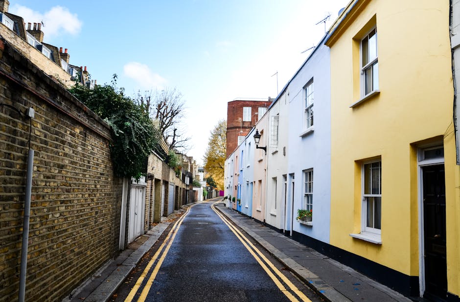 A narrow residential street with a gently curving layout, featuring a wet asphalt surface edged by a concrete sidewalk with yellow double lines along the curb. On the left, an aged brick wall, partially covered by ivy, runs parallel to the street, with small garage doors and greenery visible behind it. On the right, a row of attached houses painted in pastel shades of yellow, white, and blue, each with white-framed sash windows, some decorated with flower boxes. A black wrought-iron lamp hangs from the yellow house, indicating typical local architecture. In the background, a taller red brick building and a few leafless trees suggest a temperate climate and nearby urban infrastructure. The scene is illuminated by natural daylight, with a partly cloudy sky overhead. This setting depicts a typical street that may present access challenges for home relocation or furniture transport, which is where professional removals services like Removals Kingston upon Thames operate to assist with navigating such narrow streets during property moves.