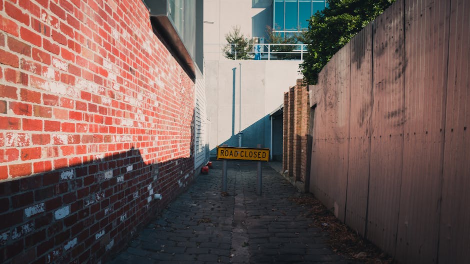 A narrow alleyway with a cobbled surface, flanked by a red brick building on the left and a tall wooden fence on the right. There is a yellow 'Road Closed' barrier positioned in the middle of the alley, indicating restricted access. The alley appears to be part of a house removal process, with no visible vehicles or furniture, but the tight space and limited access highlight potential challenges for furniture transport and home relocation activities. Bright daylight illuminates the scene, with shadows cast along the brick wall and pavement, reflecting the logistical constraints faced during moving services at Kingston Hill narrow streets as described on the page.