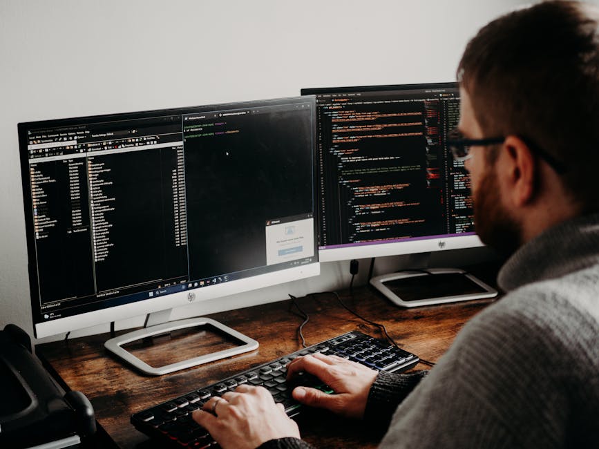 A person with a beard and glasses sits at a desk, working on a computer with two large monitors showing lines of code and data. The workspace features a wooden desk with a black telephone on the left,