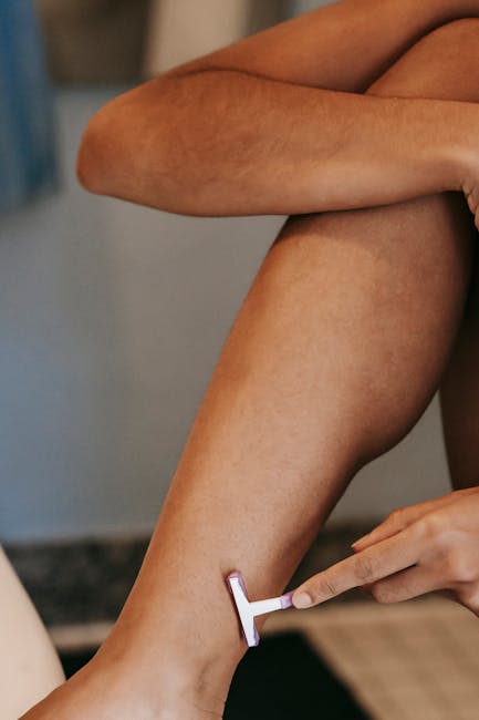A person is sitting on a surface with their legs crossed, wearing shorts, while another individual uses a razor to shave their lower leg near the ankle. The scene appears to be indoors, with a neutral background, and the focus is on the grooming activity. This image may be relevant to home relocation or packing and moving services, such as those provided by Removals Kingston upon Thames, where personal grooming is part of preparing for a move or home presentation.