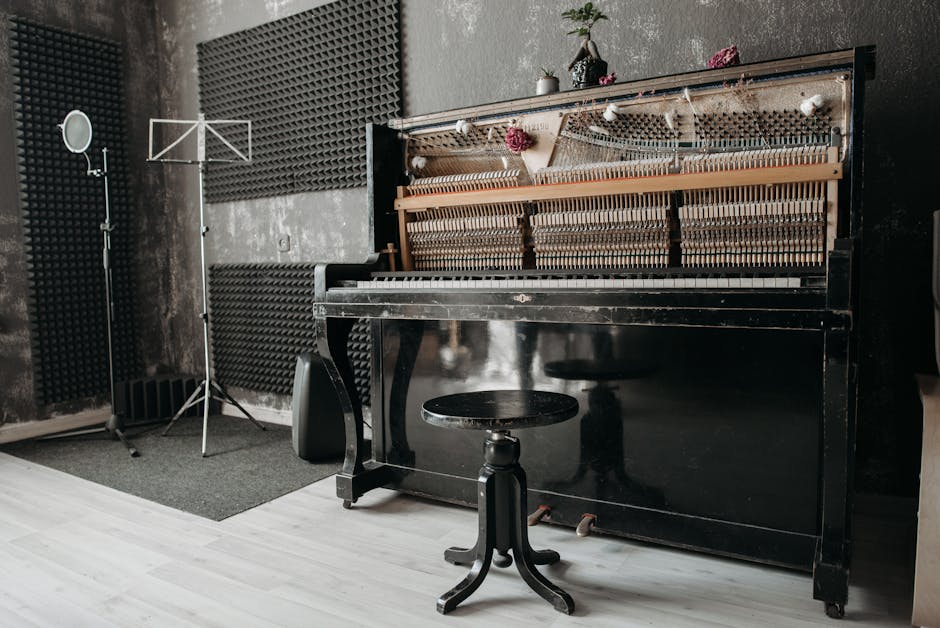 A black upright piano with its front panel removed, exposing the wooden hammers, strings, and action mechanisms, is positioned against a wall in a room with soundproofing foam panels. Several potted p