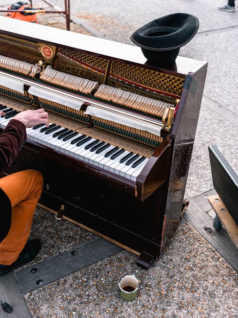 A black upright piano with its front panel removed, exposing the wooden hammers, strings, and action mechanisms, is positioned against a wall in a room with soundproofing foam panels. Several potted p