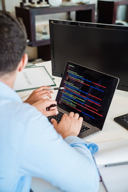 A person with dark hair, wearing a light blue shirt, seated at a desk, is working on a laptop with a black screen displaying lines of colorful code in red, blue, yellow, and green. The desk has a blac