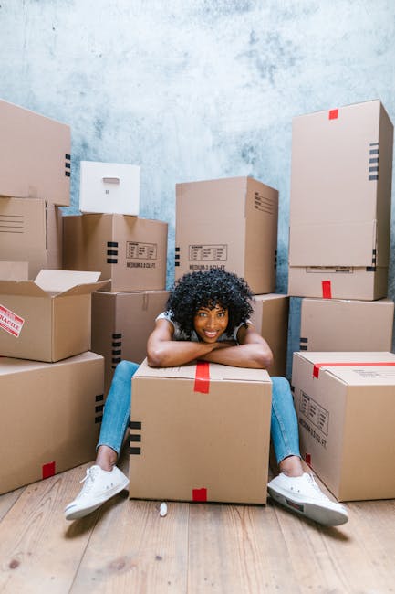 A young man with curly hair wearing a blue shirt and dark trousers is sitting on a wooden floor inside a room with large arched windows that let in natural daylight. He is surrounded by several packed cardboard boxes, some sealed with red tape, indicating a recent or ongoing move as part of a home relocation process. There is a tall potted plant with broad green leaves positioned near the window on the right side of the image. The room appears to be in a residential building with a view of parked cars and exterior buildings visible through the windows. The scene captures a moment during furniture transport or packing and moving, with the man taking a break amidst the boxes, reflective of the logistical activities carried out by professional removals, such as those offered by Removals Kingston upon Thames. The lighting highlights both the interior space and the task of loading or organizing belongings for a move.