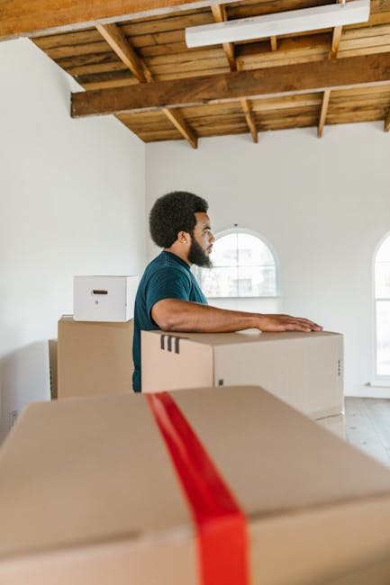 A man with dark skin and an afro hairstyle is standing inside a bright, empty room with white walls and wooden flooring. He is wearing a blue T-shirt, black trousers, and has a sweatshirt tied around